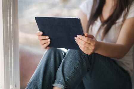 Mid selection of Attractive young woman in white medical mask sits on windowsiil and use tablet. Female have a rest at home in quarantineの写真素材