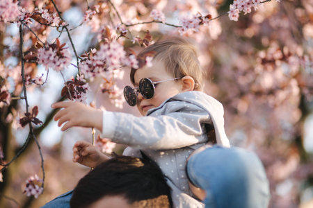 Adorable little daughter sitting on dad's neck and smels pink flowers from flowering tree.