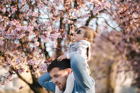 Adorable little daughter sitting on dads neck and smels pink flowers from flowering tree. Happy familyの写真素材