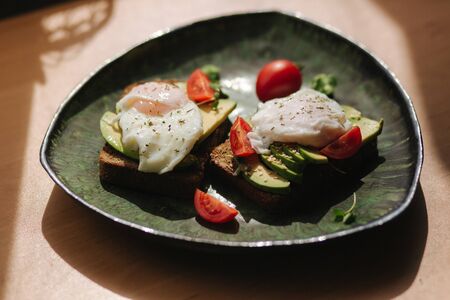Delicious breakfast at home. Sandwich with fresh sliced avocado above rye toasted bread with cherry tomatoes and poached egg on green plateの写真素材