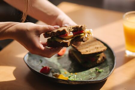 Woman hold in hands bitten sandwich. Rye toasted bread with avocado. Fresh orange juice Vegan food.の写真素材