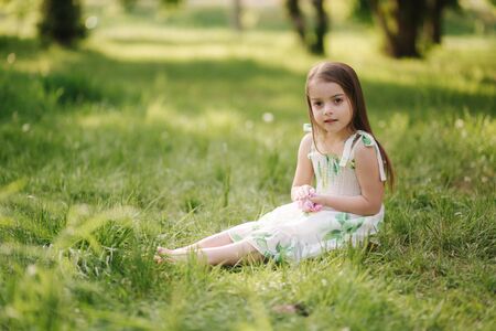 Portrait of adorable little girl sits barefoot on the grass in the park. Happy kid on the fresh airの写真素材