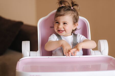 Portrait of cute baby girl sitting in high chair in the kitchen. Adorable little girl waiting for dinnerの写真素材