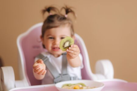 Beautiful little baby girl first time eating exotic fruits in high chair. Cute baby girl taste delitious fruist, mango, kiwi, and babanaの写真素材