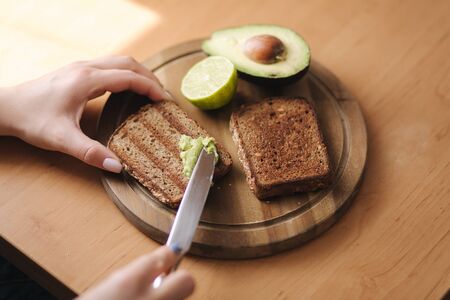 Woman puts guacamole or avocado spread on top of rye bread toast on wooden board at home. Vegan breakfastの写真素材