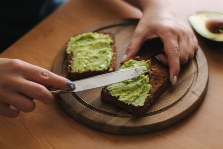 Close-up of Woman puts guacamole or avocado spread on top of rye bread toast on wooden board at home. Vegan breakfastの写真素材