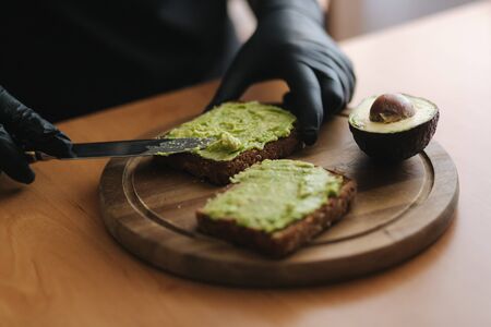 Close-up of Woman in black glowes puts guacamole or avocado spread on top of rye bread toast on wooden board at home. Vegan breakfastの写真素材