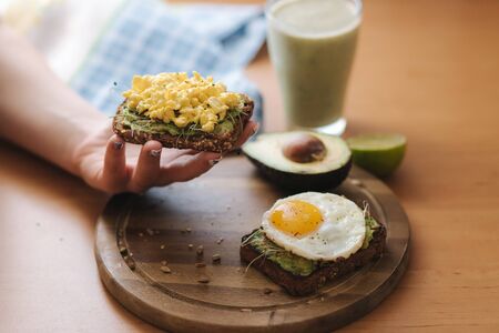 Woman hold toasted sandwich. Egg on avocado sandwich with whole grain bread on wooden board. Smoothie with spinachの写真素材