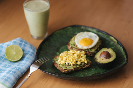 Different cooked egg on avocado sandwich with whole grain bread on wooden board. Smoothie with...