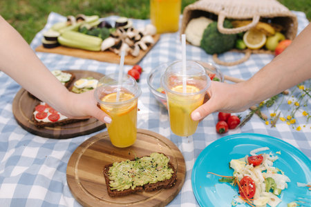 Photo of two hand hold orange lemonade in front of vegan picnic outdoors. Heathy food concept.