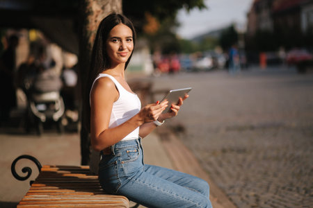 Beautiful girl use tablet outdoors. Female sitting on the bench alone. Woman watch video on tablet...