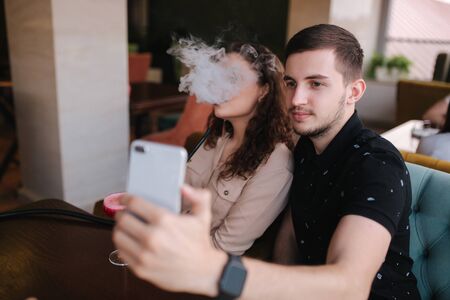 Group of people smoke hookah on terrace. Man and women meet in restaurant. Man make stories while his girlfriend smoke. Man make selfieの写真素材