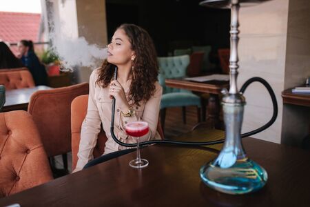 Attractive young woman smoke hookah in restaurant. Woman with hookah on terrace by herselfの写真素材