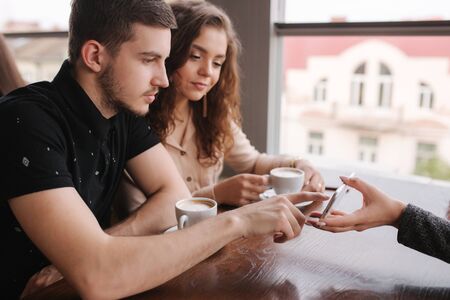 Happy couple in cafe look into smartphone and smile. Woman show to man and woman something in phone. Man put finger on screen and smileの写真素材