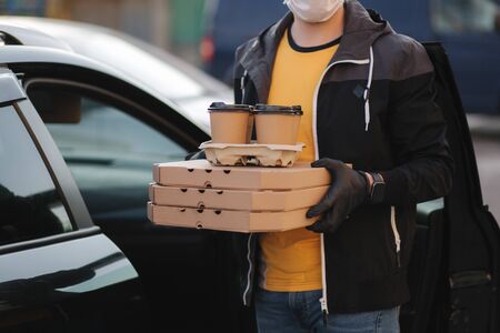Courier in protective mask and medical gloves take order from car. Delivery boy hold pizza and coffe. Delivery service under quarantine. Coronavirus covid-19 themeの写真素材