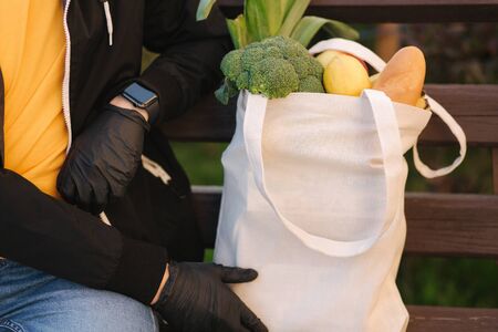 Courier in protective mask and medical gloves delivers fresh food. Delivery boy sit on bench and wait for customer. Delivery service under quarantine. Coronavirus covid-19 theme. Middle selection.の写真素材
