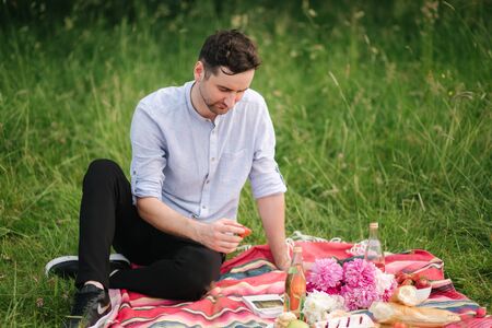 Handsome man sitting on red blanket outdoors and waiting his girlfriend for picnicの写真素材