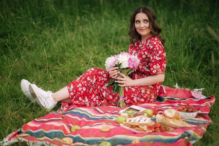 Young woman in red dress sits on red blanket outdoors. Female hold flowers. Mini picnincの写真素材
