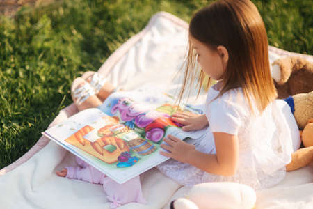 Adorable little girl sits on plaid outdoors and read book. Happy smiled baby girl spend time on back yard in sunsetの写真素材