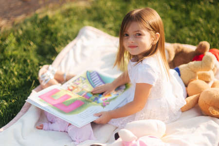 Adorable little girl sits on plaid outdoors and read book. Happy smiled baby girl spend time on back yard in sunsetの写真素材