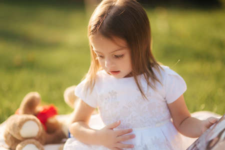 Close up of Adorable little girl sits on plaid outdoors and read book. Happy smiled baby girl spend time on back yard in sunsetの写真素材