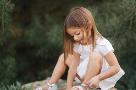 Little girl in white dress sits on big stone in front of green conifer treeの写真素材