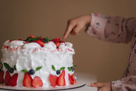 Little girl first time taste bithday cake. Cute little girl in beautiful dress tasting cake with strawberries. Close up of girl put finger into cakeの写真素材