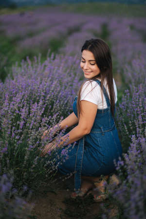 Young woman in denim dress sitting in lavender field in summer. Beautiful girl smileの写真素材
