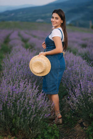 Attractive young woman walking in summer lavender field. Model dressed in denim sundress with straw hat and bagの写真素材