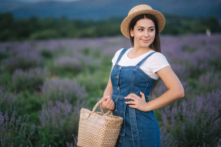 Portrait of attractive woman in lavender field in straw hat and denim sundress. Farm style in lavender with straw bagの写真素材