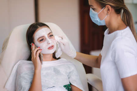 Hands of cosmetology specialist removing white facial mask using brush. Making skin hydrated and face glowing and skin. Hulf face with clay mask. Attractive young brunette relaxing with closed eyes and enjoying spa procedures. Close up.の写真素材