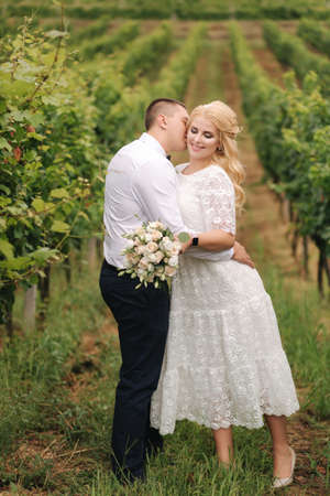 Groom and bride walking in vineyard in their wedding day. Blond hair woman in white dress hold wedding bouquet in handsの写真素材