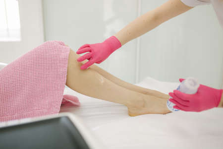 Young woman beautician in mask applies contact gel on the leg of a woman for laser hair removal in medical clinic. Close up. Pink glowesの写真素材