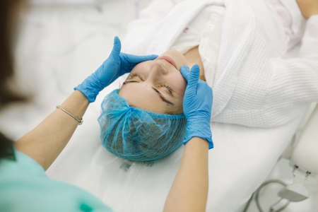 Cosmetoligist make facial massage in beauty salon. Preparing for injection. Cosmetologist in protective medical maskの写真素材