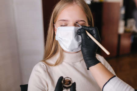 Brow master in black protective mask applies henna on eyebrows for beautiful blond hair woman. Customer in medical mask. First person viewの写真素材