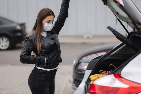 Young stylish woman in protective mask with shopping cart full of fresh food during quarantine,...