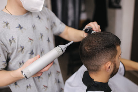 Hairdresser use hairdryer for making a hairdress, Barber and client in face mask