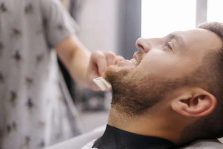 Professional hairdresser uses a hair clipper for fringing beard for a handsome man in barbershop. side viewの写真素材