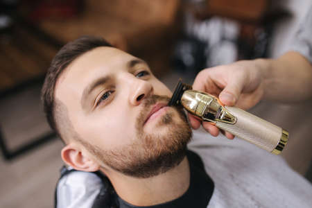 Professional hairdresser uses a hair clipper for fringing beard for a handsome man in barbershopの写真素材