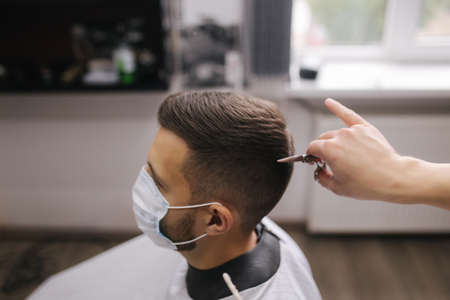 Barber cutting hair to a handsome bearded man in face mask. Protective mask on client face. The hairdresser also wears a mask. Quarantine haircut concept.の写真素材