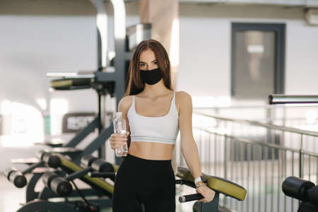 Attractive brunette female have a break in gym. Woman in protective mask and hold bottle of waterの写真素材