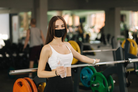 Attractive brunette female have a break in gym. Woman in protective mask and hold bottle of water