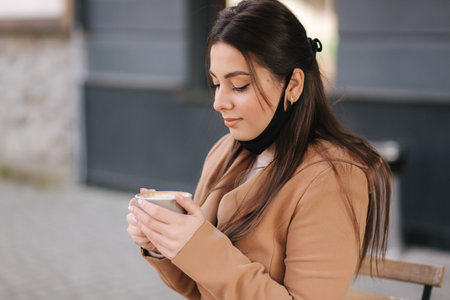 Portraitf of Beautiful woman takes off protective mask for drinking coffee. Female sitting outdoors...