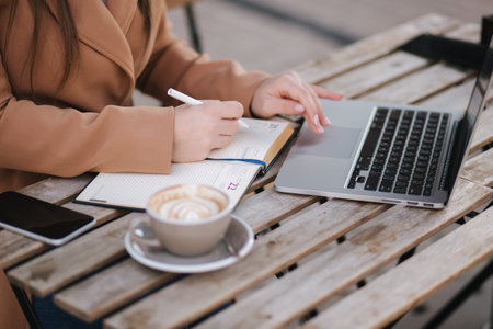 Close up of working proces in cafe outdoors. Woman writing down something in notebook. Cappuccino...