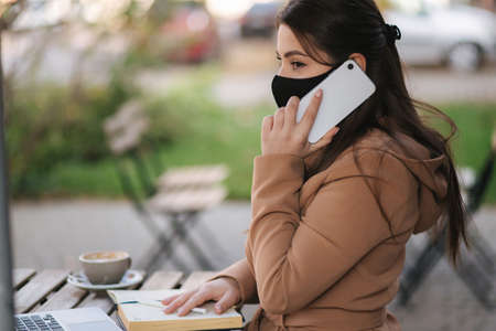 Young female in black face mask sitting on terrace in cafe during quarantine. Business woman working outdoos in quarantine. Woman speak by the phone. Covid-19の写真素材