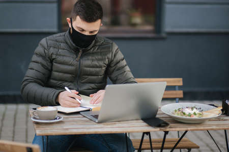 Handsome man in face mask sitting outdoors in cafe and working on laptop. Man take salad. Quarantine. Covid-19の写真素材