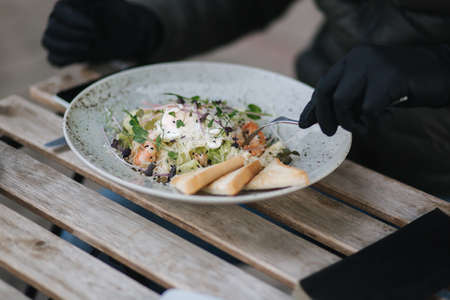 Close up of man in black gloves hold cutlery. Cafe concept during qurantine. Food outdoors in protective gloves. Fresh vegan salad with shrimpsの写真素材