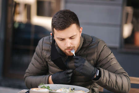 Man in black gloves hold cutlery. Quarantine Cafe concept. Food outdoors in protective gloves. Fresh vegan salad with shrimps. Man pulled off protective mask for eatingの写真素材