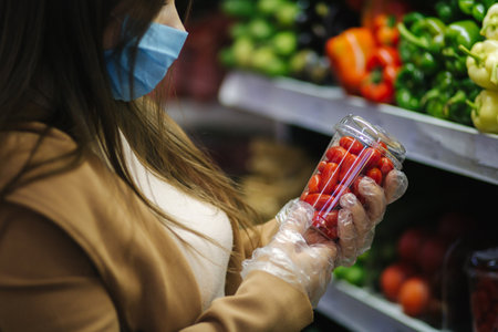 Happy woman taking fresh cherry tomatoes while standing at groceries in supermarket. Beautiful young girl choosing red tomatoes by stand with vegetables.の写真素材