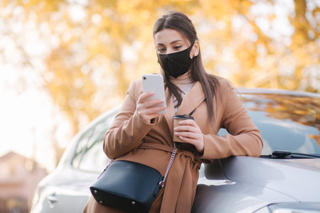 Attractive young woman in protective mask with cup of coffee stand by the car and use phone. Cafe is closed woman drink coffee outdoors safetyの写真素材